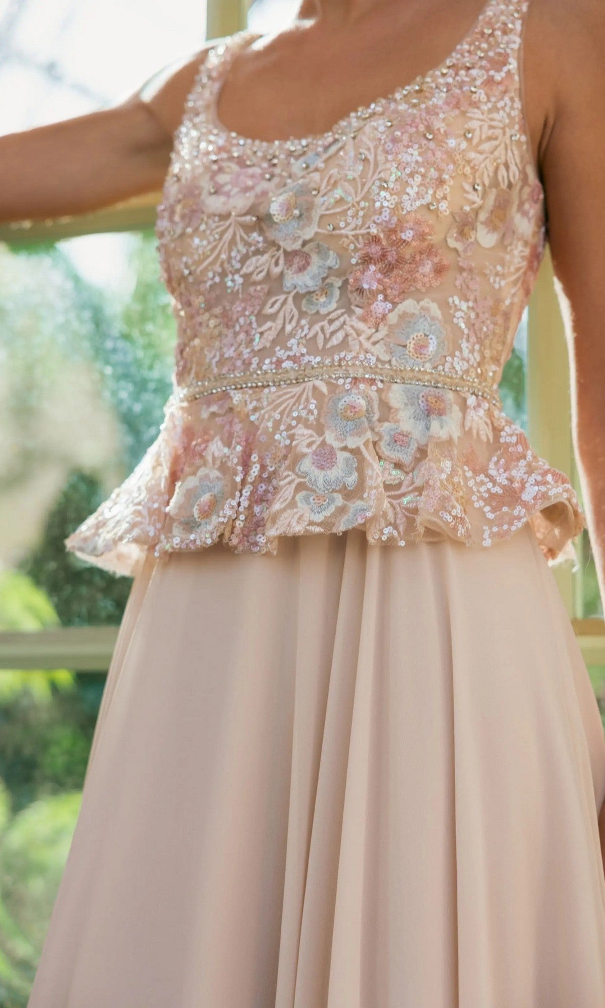 A woman wearing the Long Formal Dress: Mac Duggal 21317โa sleeveless blush pink A-line chiffon evening gown featuring a floral embroidered bodice and a peplum waistlineโstands near a sunlit window.